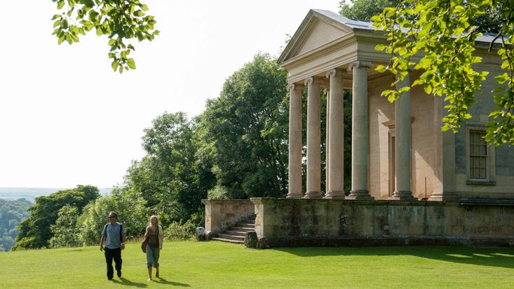 Two visitors walking on the lawn close to the grand Ionic Temple at Rievaulx Terrace, North Yorkshire, on a sunny day
